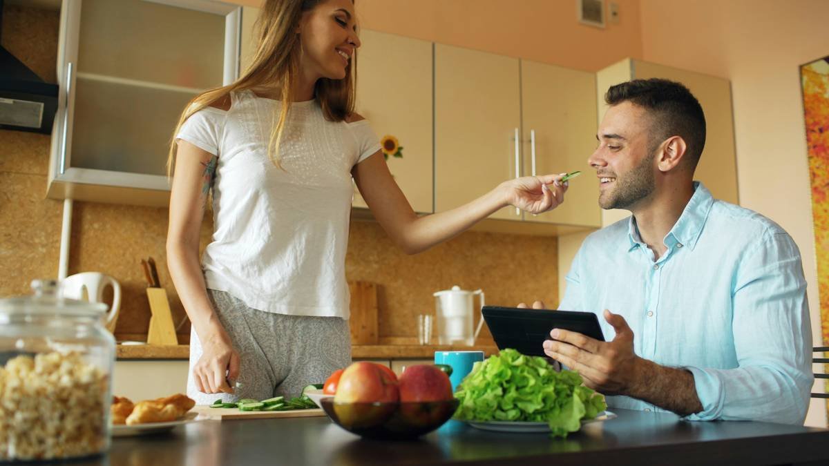 Attractive couple chatting in the kitchen early morning. Beautiful woman feed her husband while cooking breakfast at home